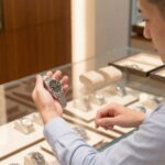 高級時計店のガラスショーケースの前で、ステンレス製のロレックス・デイトジャスト（黒文字盤）を左手で持ち、真剣な表情で見つめる日本人男性。ショーケース内には他のモデルも並んでおり、比較検討している様子。